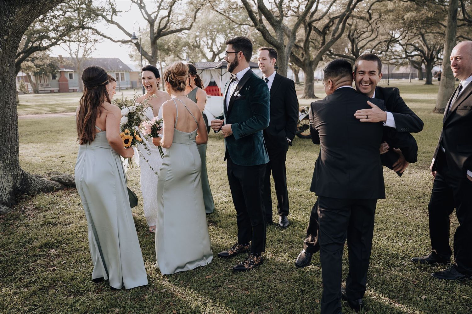 Wedding party mingles casually outdoors under trees during celebration.