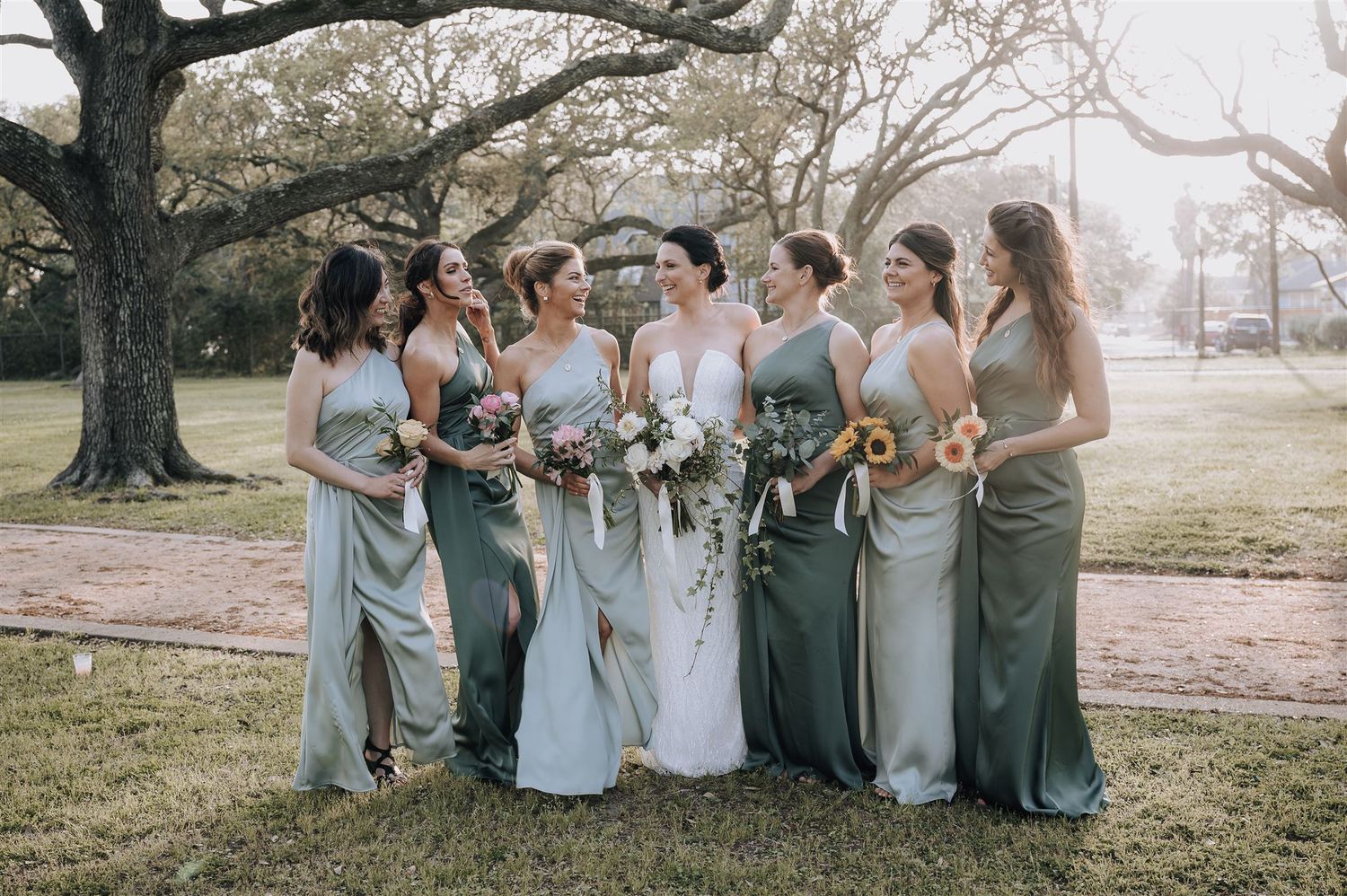 Bridal party in varying shades of sage green dresses pose together under blooming trees.