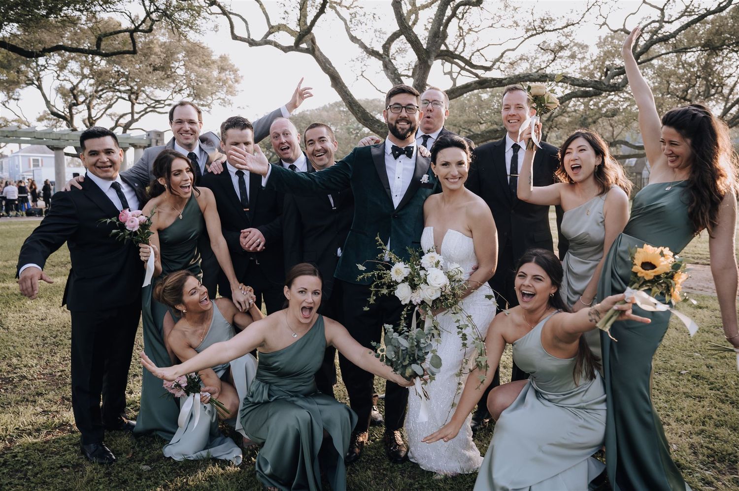 Wedding party celebrates joyfully outdoors with bridesmaids in sage green dresses and groomsmen in black tuxedos.