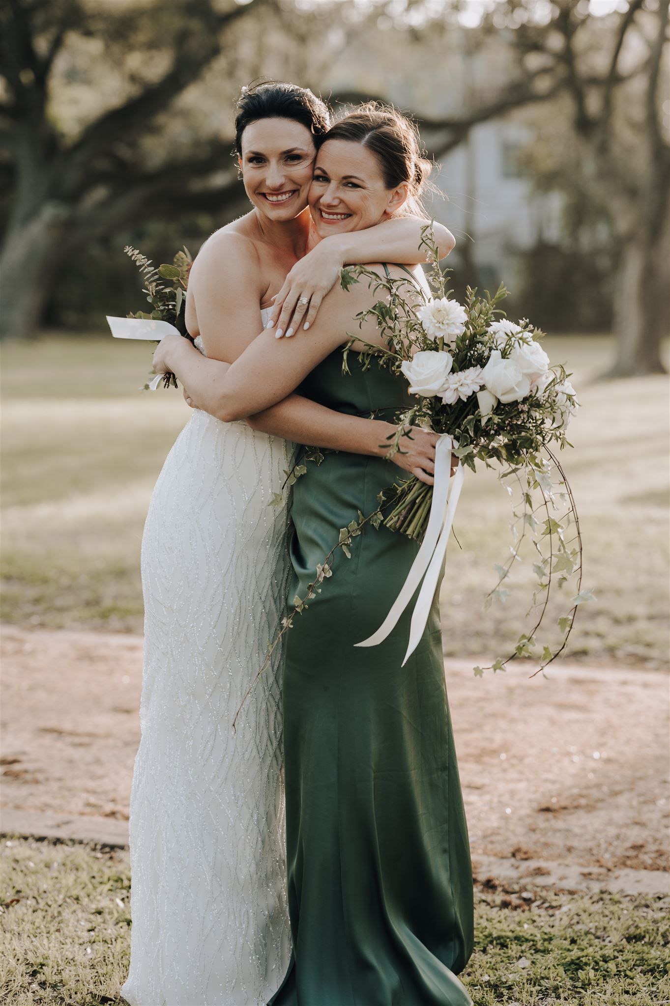 Tender embrace between bridesmaids wearing white and emerald green dresses with cascading bouquets.