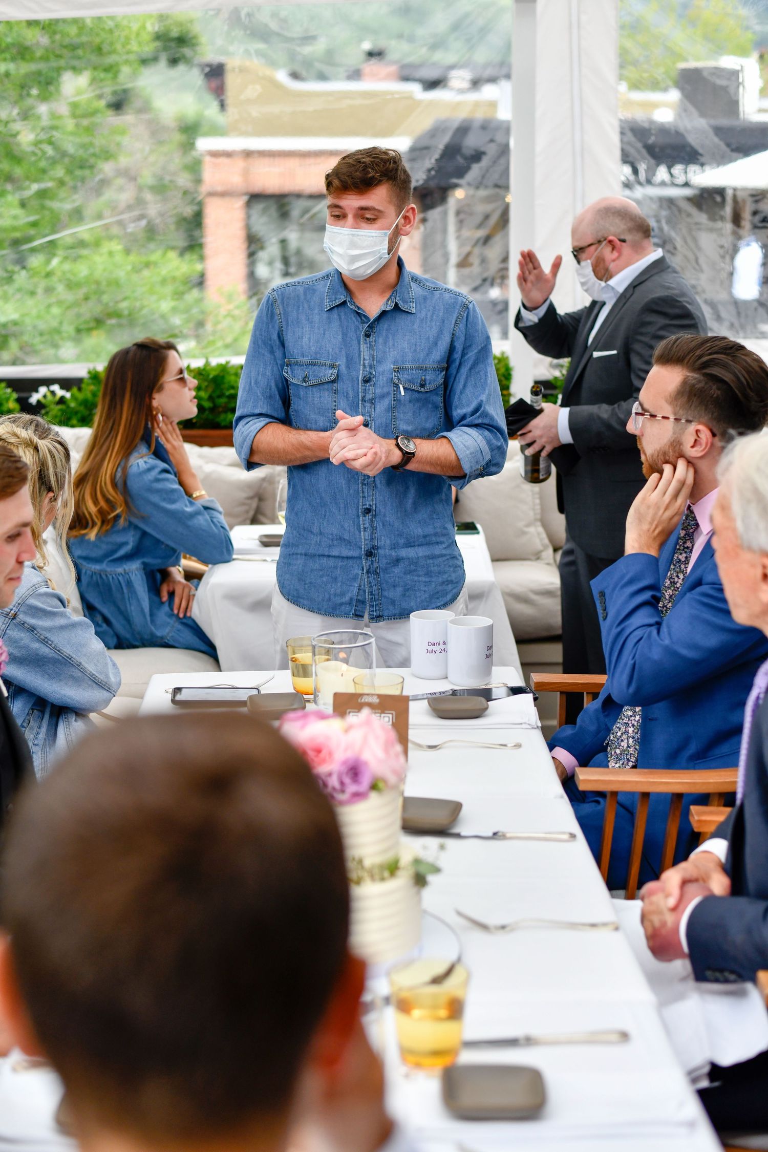 People gathered at an outdoor dining table wearing casual attire and face masks.