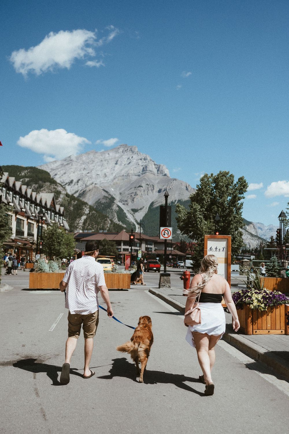 People walking with dog on mountain town street with scenic peaks in background.