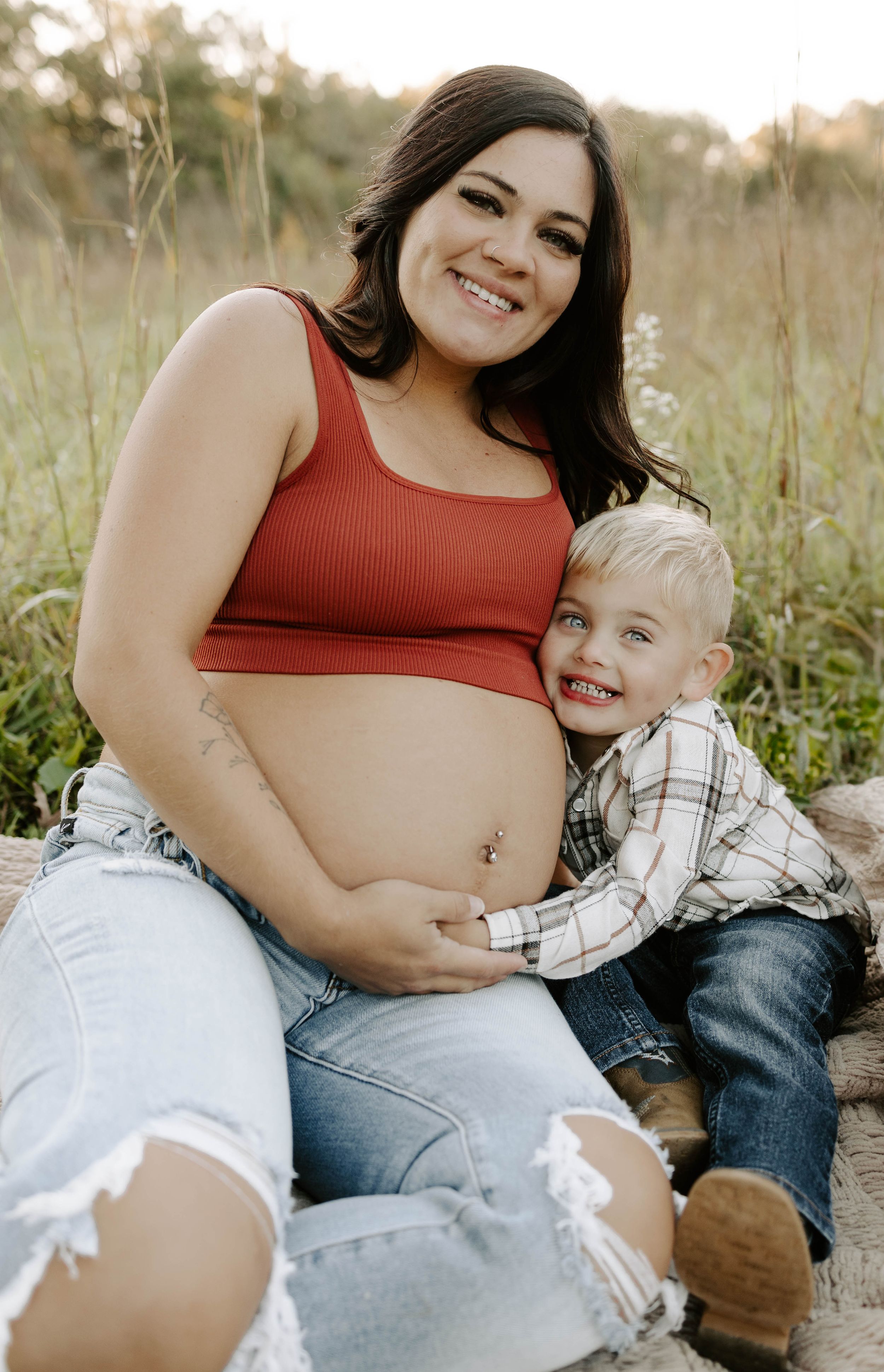 Joyful maternity photo with young child hugging mother's baby bump in grassy field.