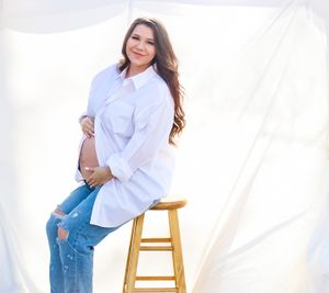 Woman in white button-down shirt and distressed jeans sitting on wooden stool against white backdrop.
