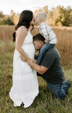 Sweet family maternity photo with child and father kissing pregnant mom's belly in golden field.