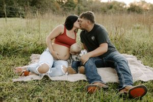 Expecting couple shares intimate moment on picnic blanket in rustic outdoor setting.