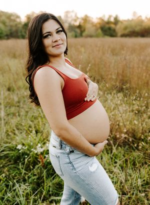 An expectant mother in a red crop top and jeans poses in a sunlit field.
