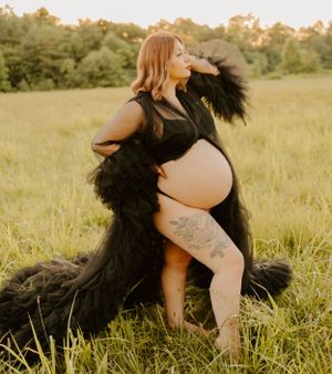 Artistic maternity photo featuring a woman in black dress with feathers posing in golden field at sunset.