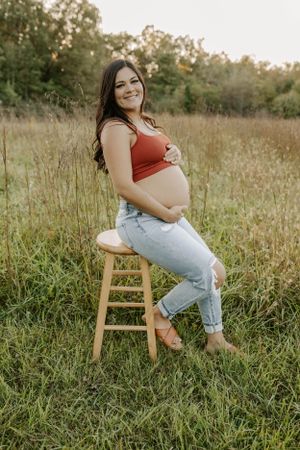 Pregnant woman in red top and jeans sits confidently on wooden stool in grassy field.