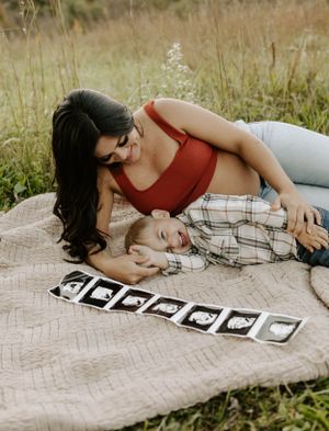 A maternity photoshoot features ultrasound photos laid out on a blanket in a grassy field.