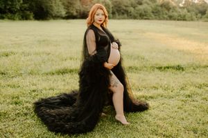 Expectant mother in flowing black lace gown poses gracefully in sunlit meadow.