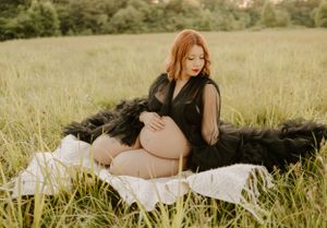 Maternity portrait of an expectant mother in a black sheer dress reclining on white blankets in a sunlit meadow.