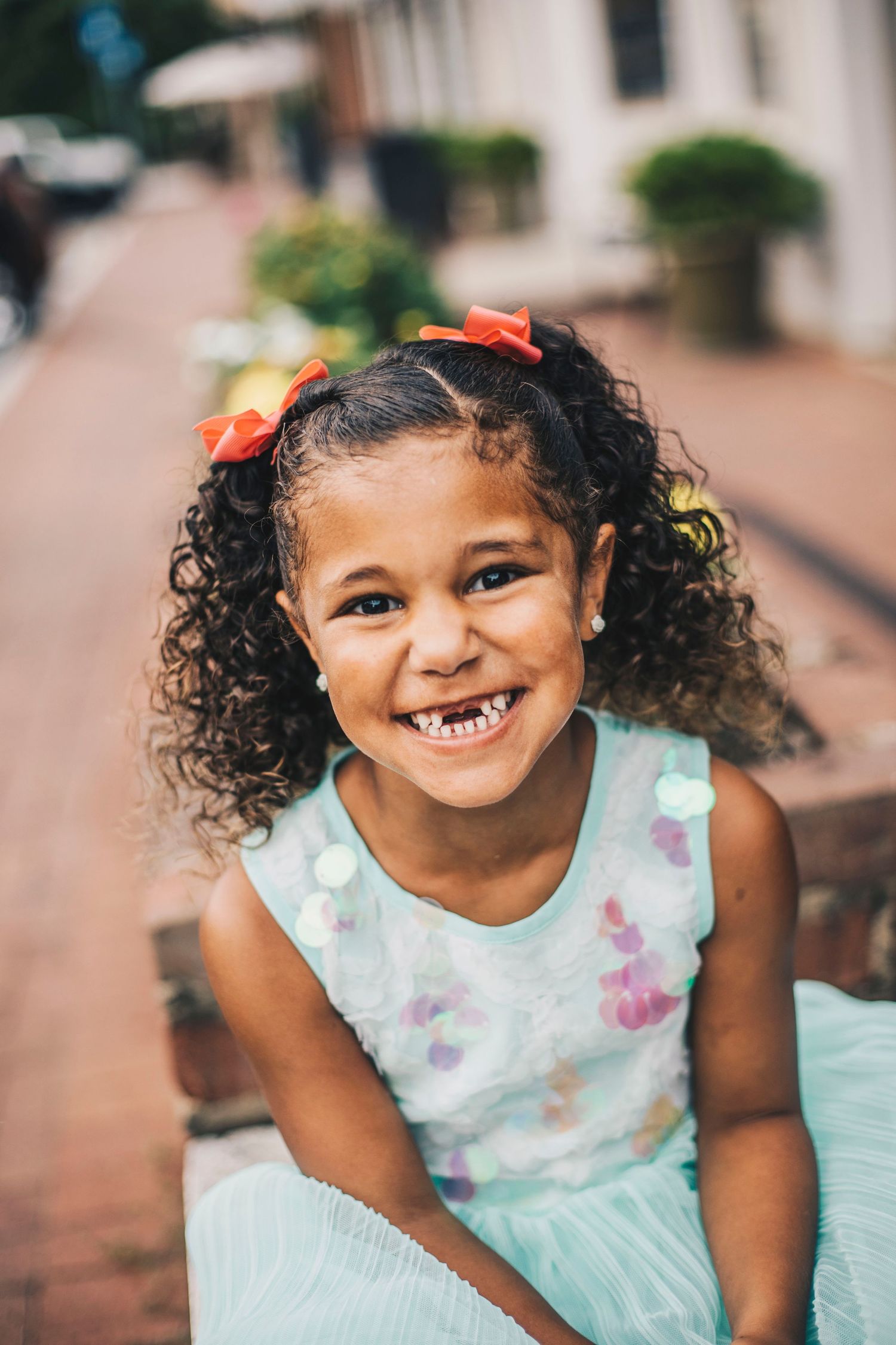 Smiling young girl with curly hair and red bows wearing a mint sequin dress outdoors.