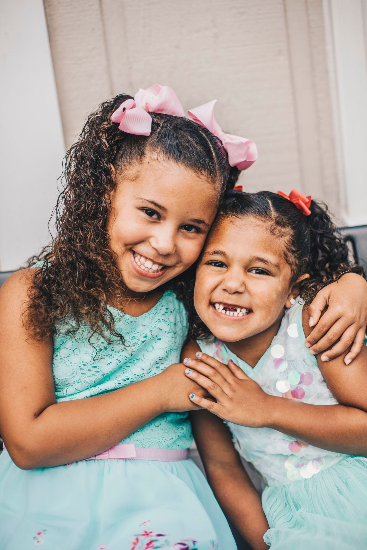 Two smiling sisters with curly hair in mint dresses hugging each other closely.