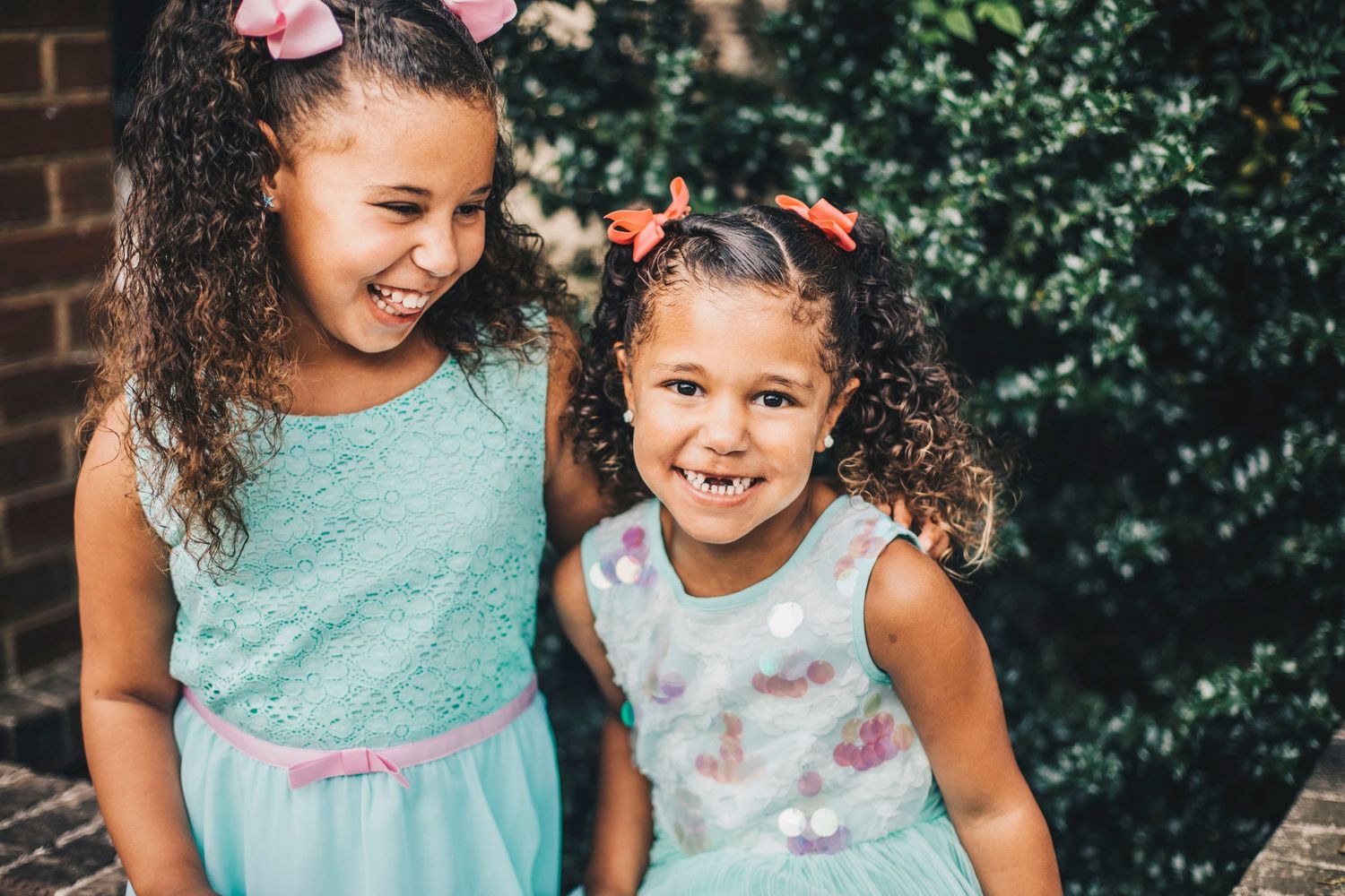 Two young girls in mint dresses smiling together outdoors near green bushes.