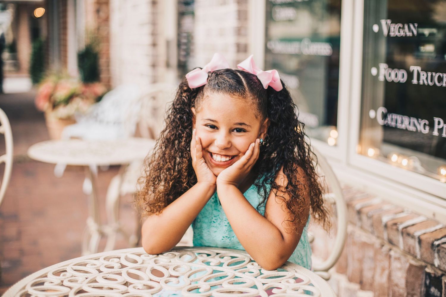 Young girl in mint dress resting chin on hands at white ornate outdoor cafe table.