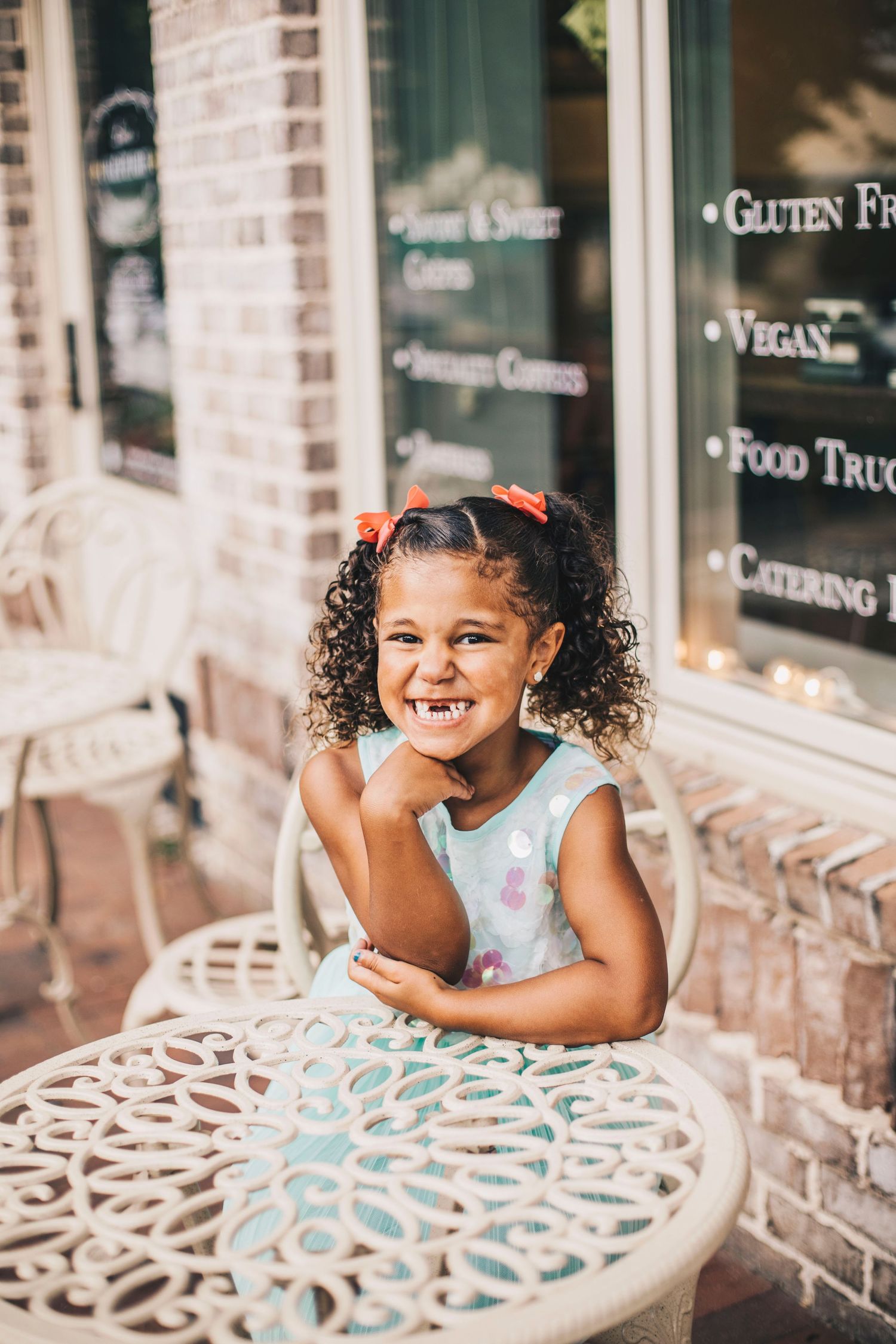 Young curly-haired girl in mint dress sitting at white ornate cafe table outdoors.