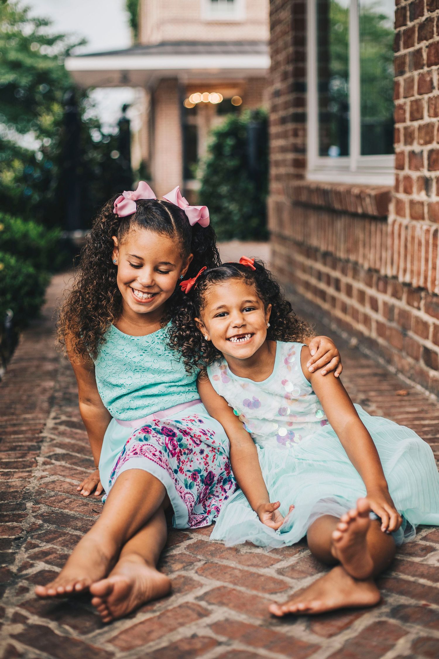 Two sisters in mint dresses sitting together on brick pathway beside brick wall.