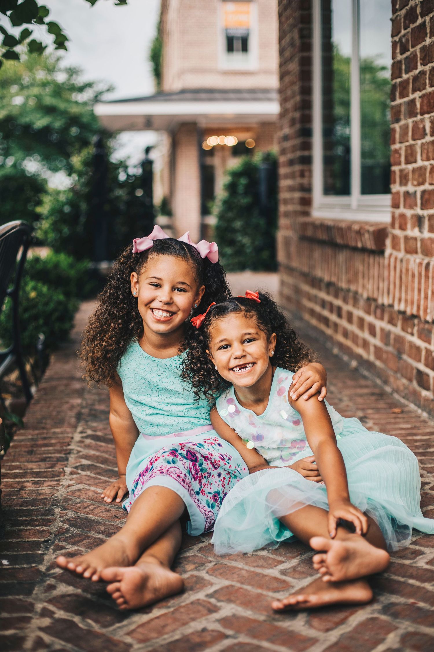 Two smiling girls in mint dresses sitting together on brick pathway near brick building.
