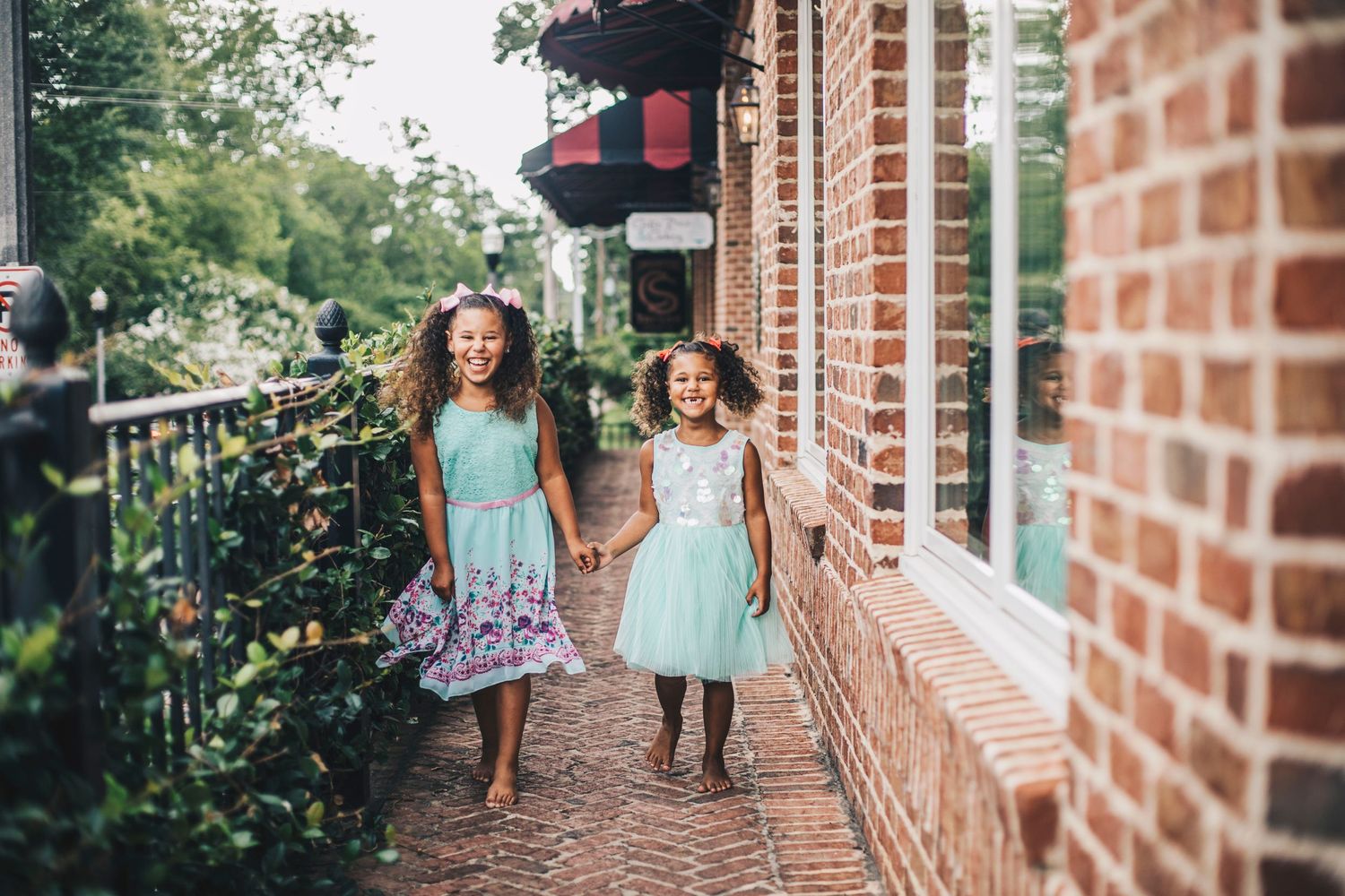 Two young girls in mint dresses holding hands walking along brick pathway outdoors.