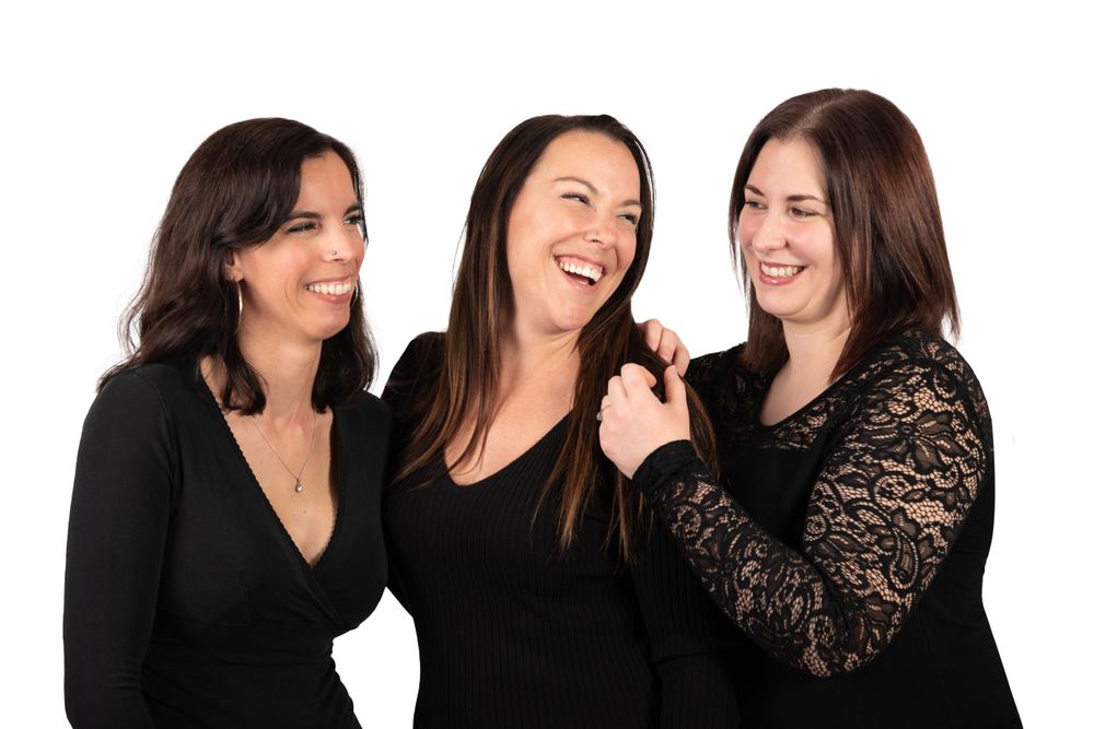 Group portrait of women in black attire laughing together against white background.