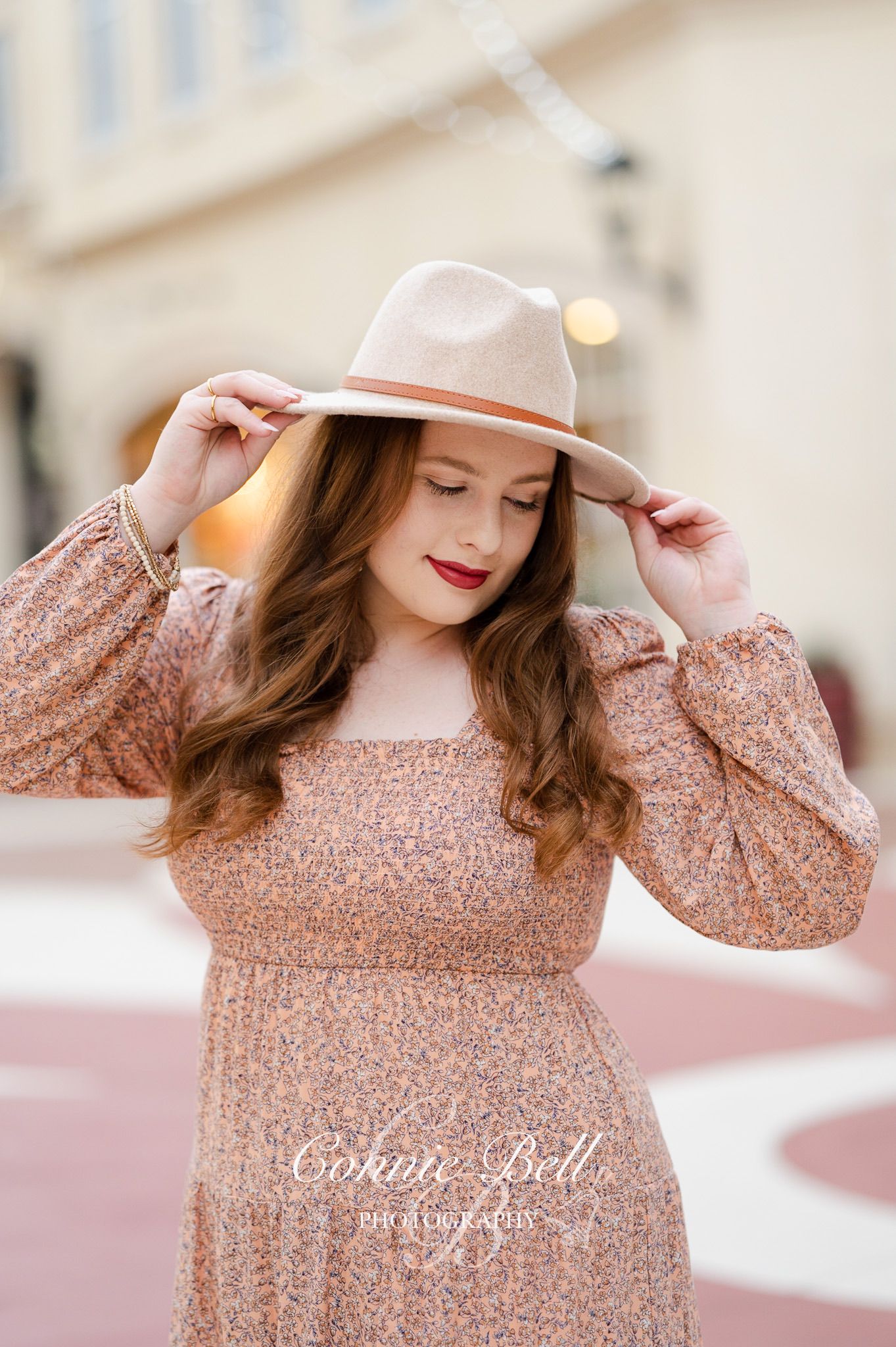 Woman in a peach floral dress and beige fedora hat poses outdoors with natural lighting and soft bokeh background.