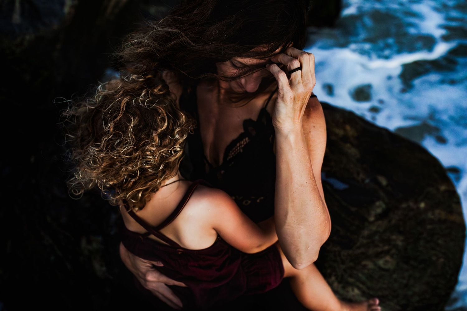 Person with curly hair sitting on dark rocks near blue water with hands covering their face.
