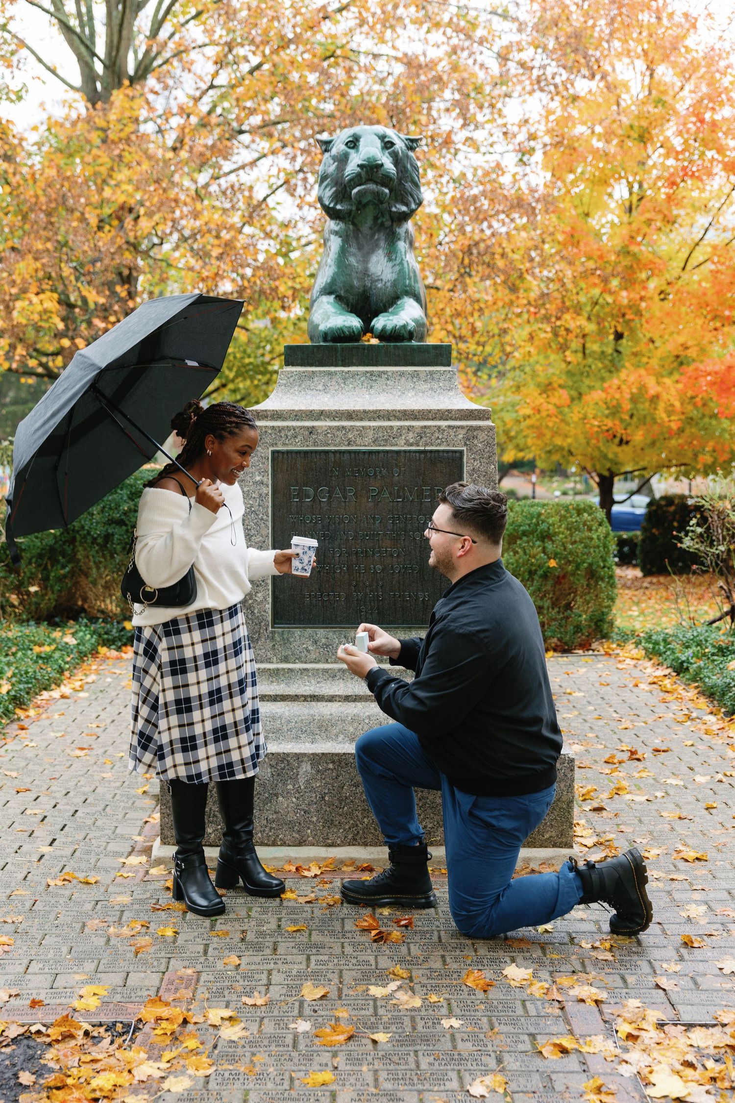 A proposal scene with umbrella near a bronze bulldog statue surrounded by fall foliage.