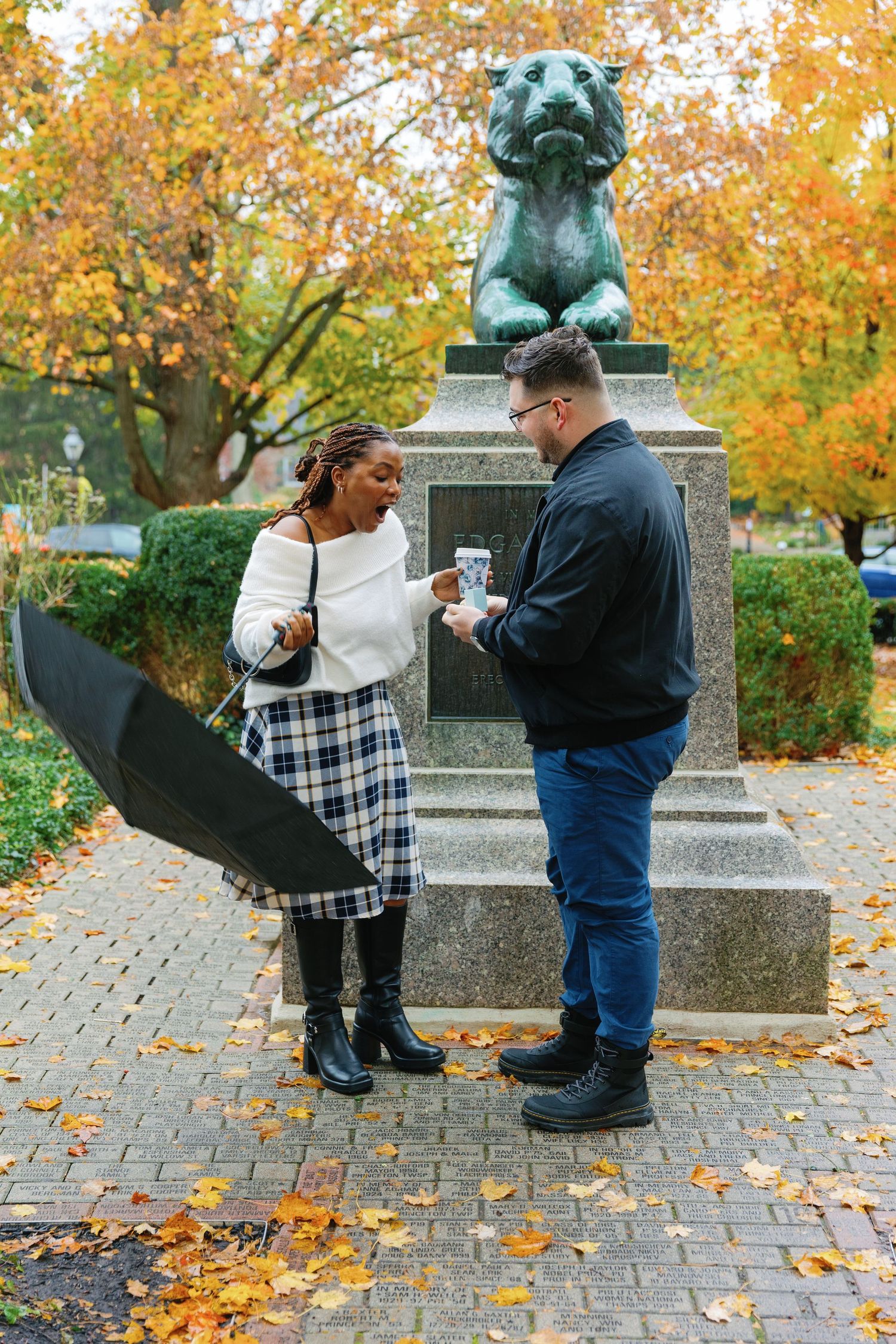 A couple during a surprise proposal by a bronze statue on leaf-covered ground.