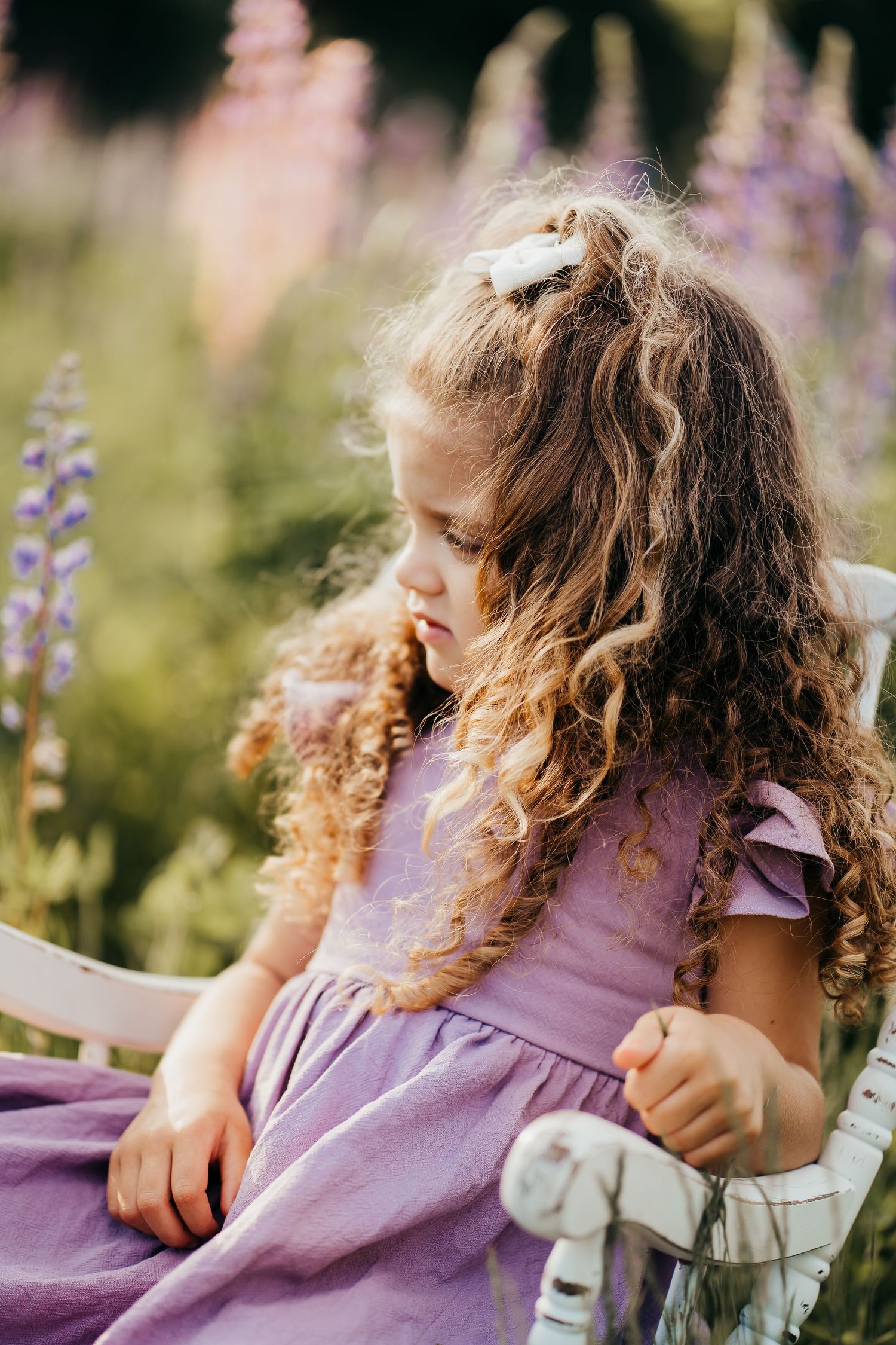 Curly-haired girl in a flowing lavender dress sits among blooming lupine flowers in a dreamy garden setting.
