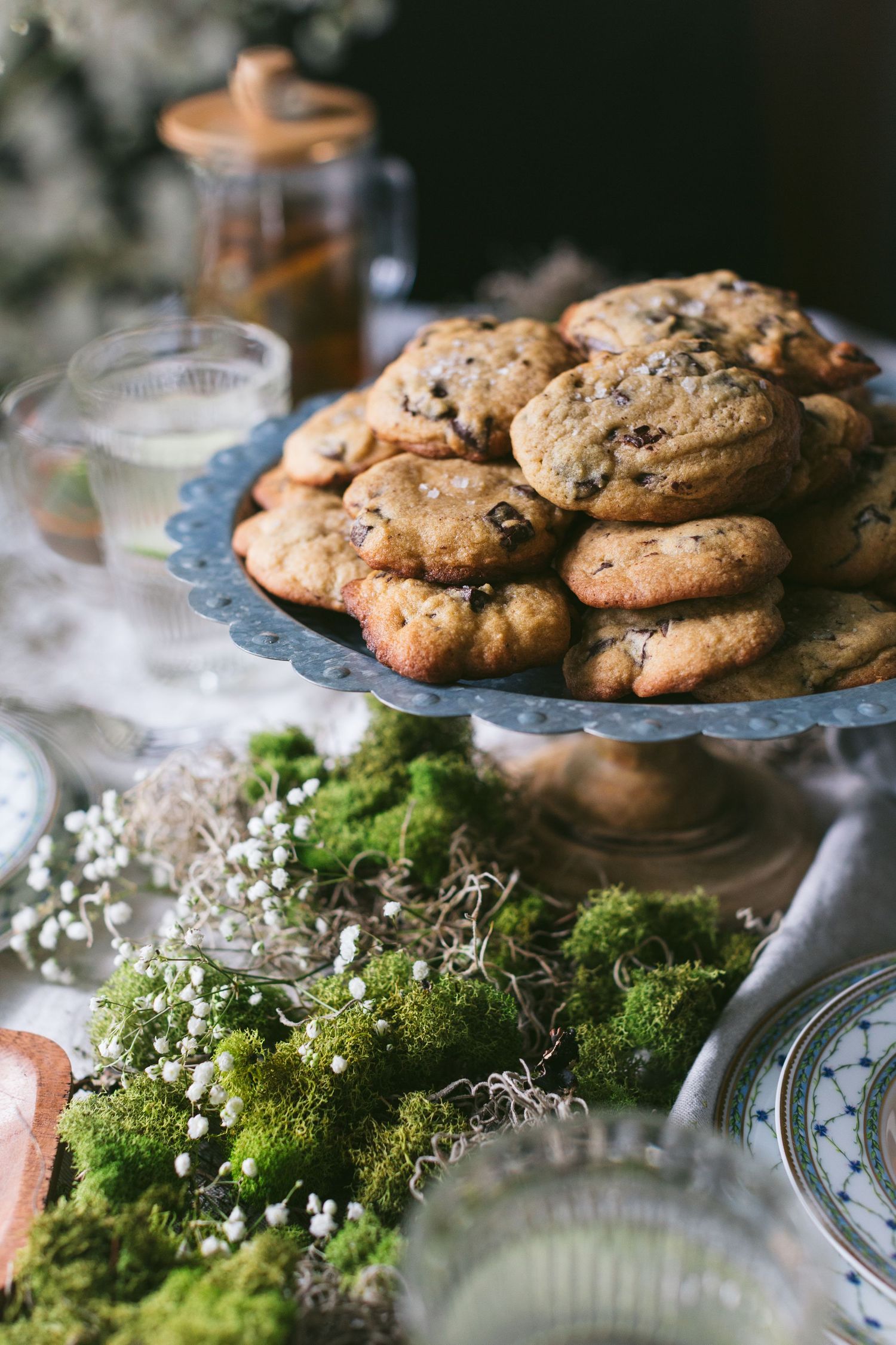 Rustic cake stand piled with chocolate chip cookies surrounded by green moss and delicate white flowers on a linen tablecloth.
