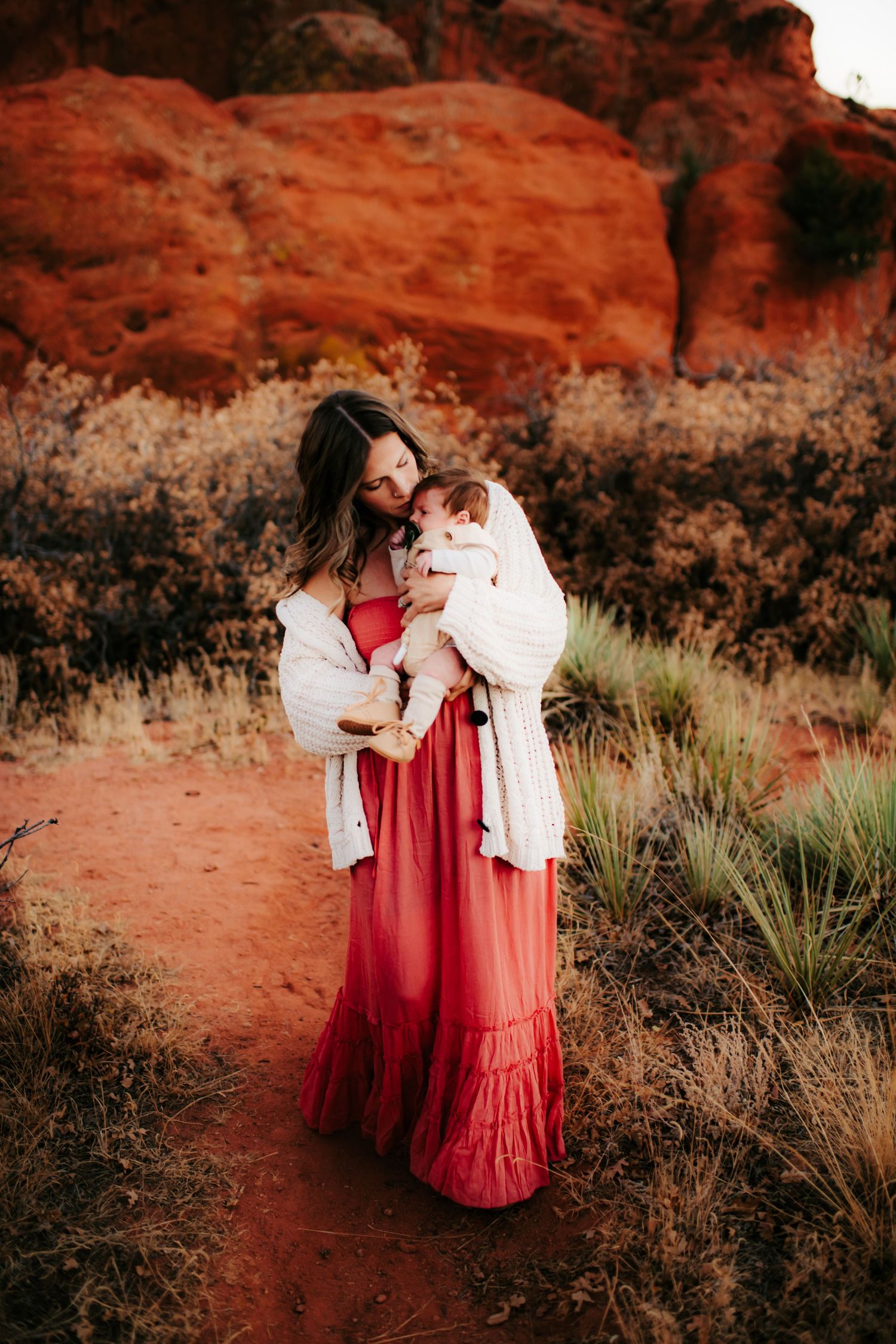 Woman in flowing red dress holding baby against backdrop of red rock formations.