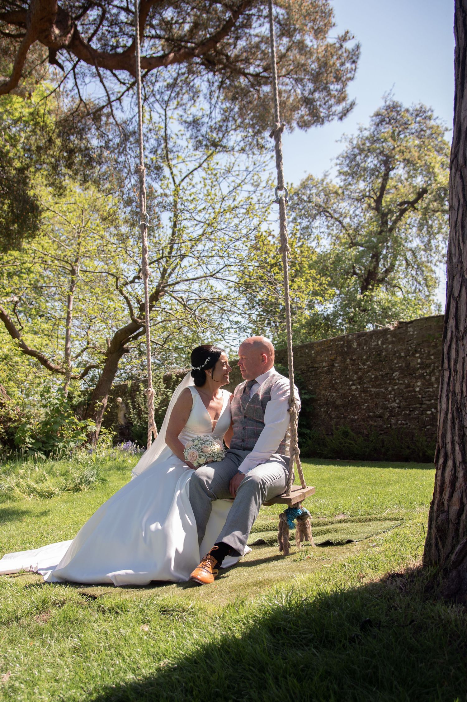 A couple in wedding attire relaxes together on a grassy slope near a stone wall on a sunny day.