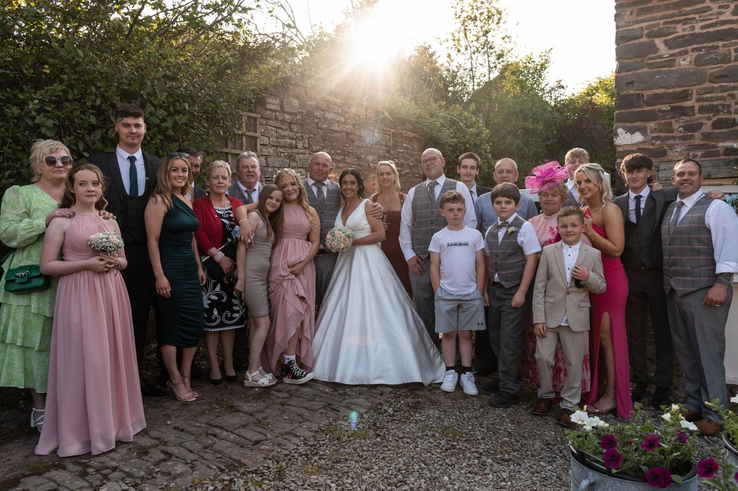 Large group wedding photo of guests gathered outside a historic stone building at sunset.