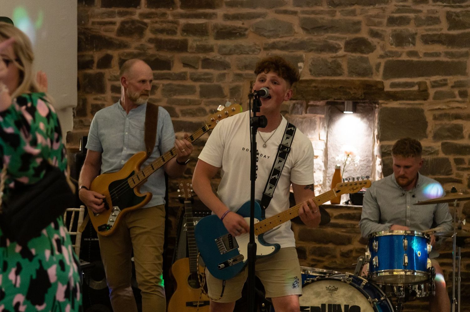 Musicians playing instruments on stage with atmospheric lighting and stone wall backdrop.