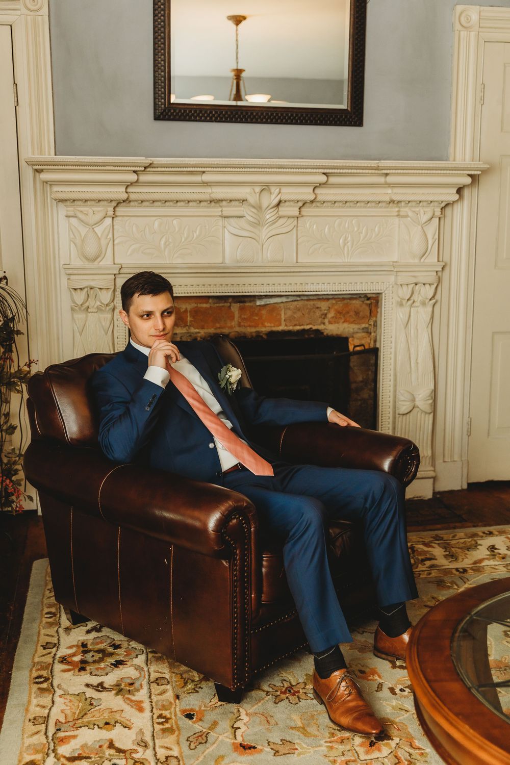 Man in blue suit sits thoughtfully in leather chair by ornate white fireplace.