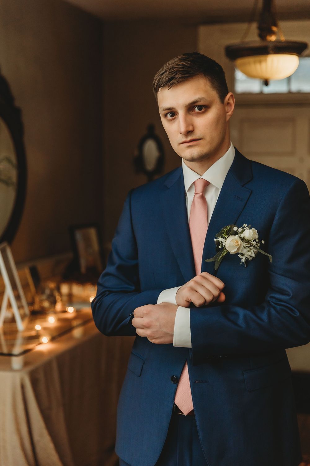 Groom in navy suit with pink tie and boutonniere standing in warmly lit room.