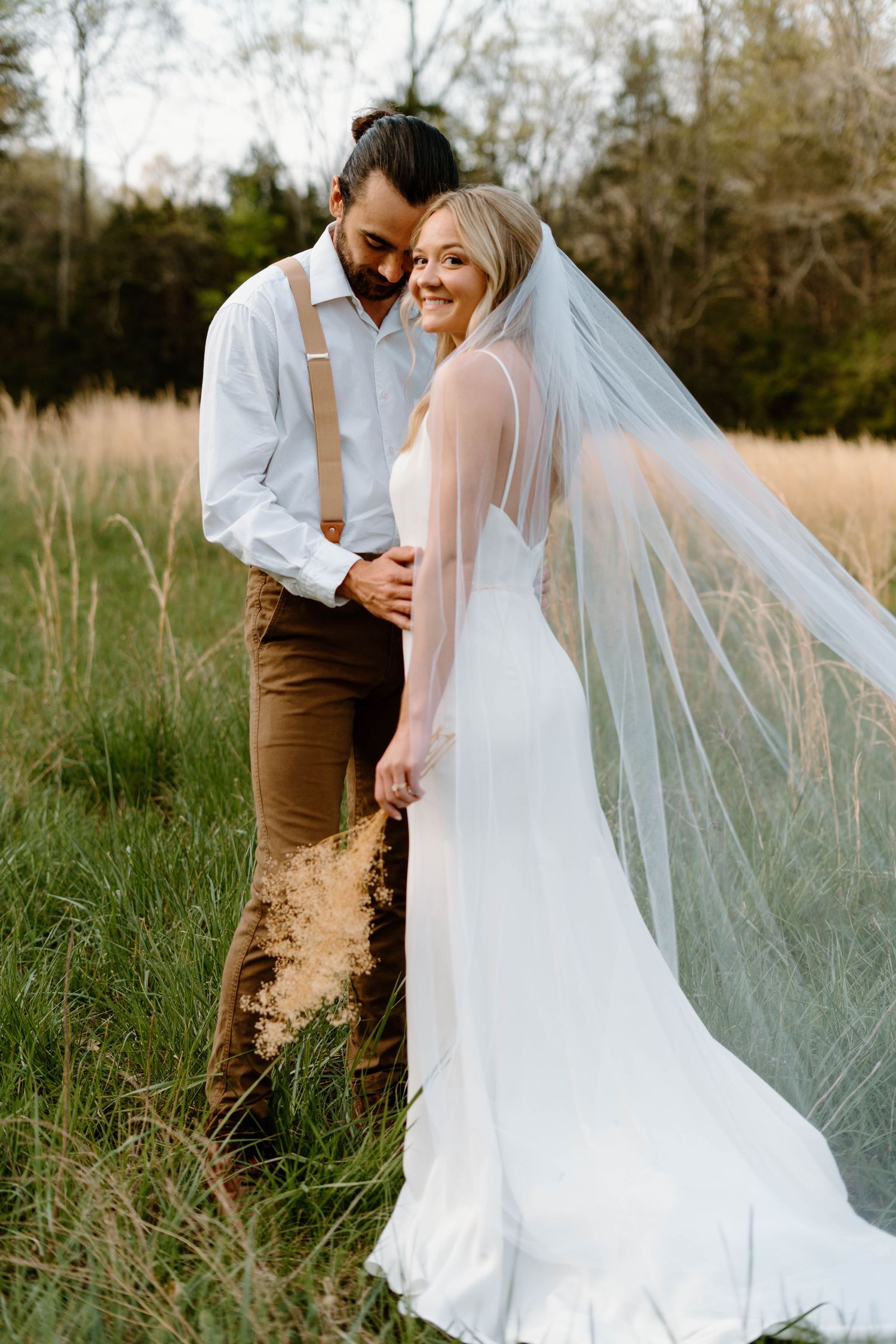 a bride with a flowing veil and groom pose in wedding attire in an open field