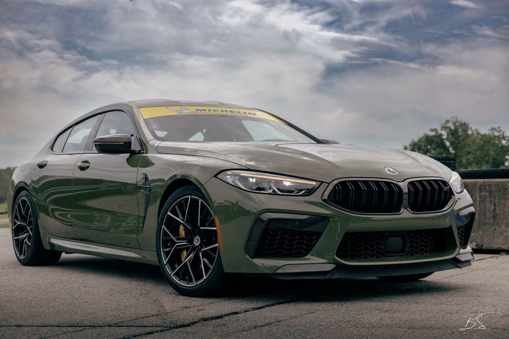 A dark green BMW M8 Gran Coupe parked on a race track under a dramatic cloudy sky.