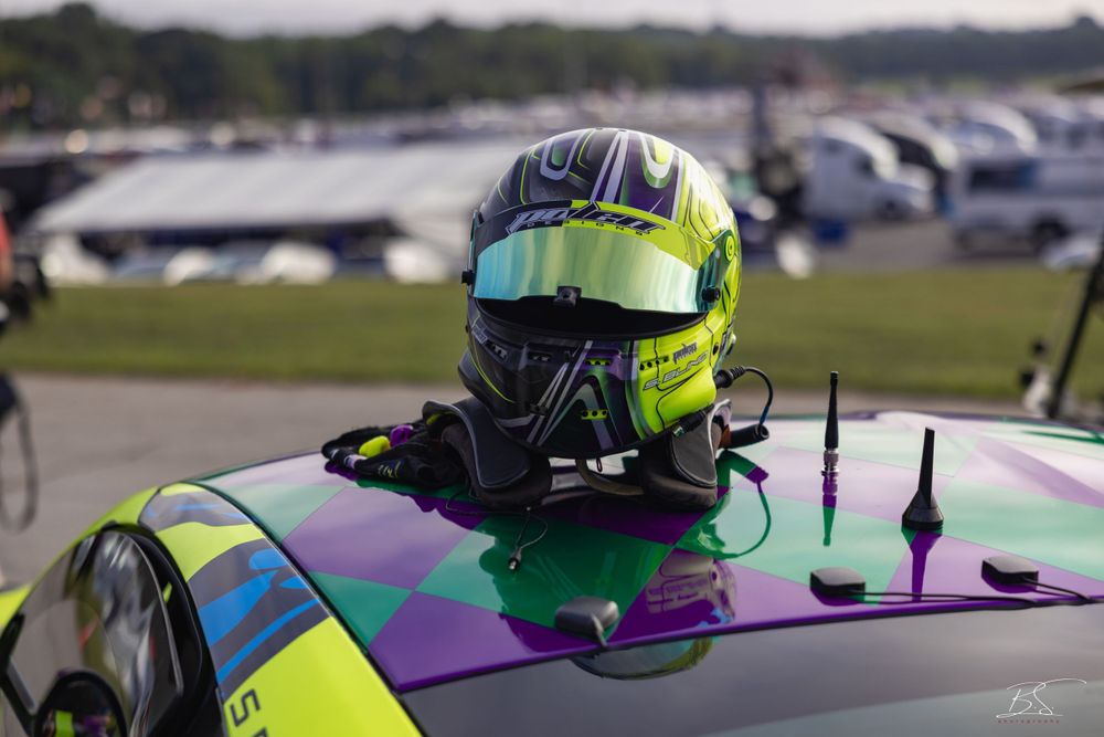 A colorful racing helmet in green and purple rests on the roof of a race car at a motorsport event.