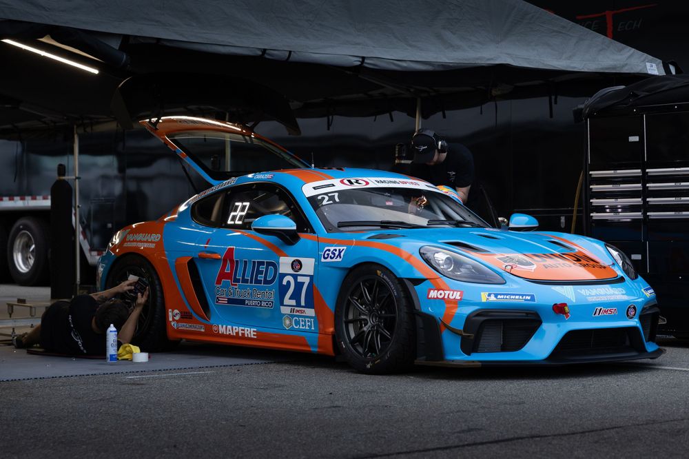 A blue and orange Gulf-liveried Porsche 718 Cayman GT4 race car number 27 in a pit garage.