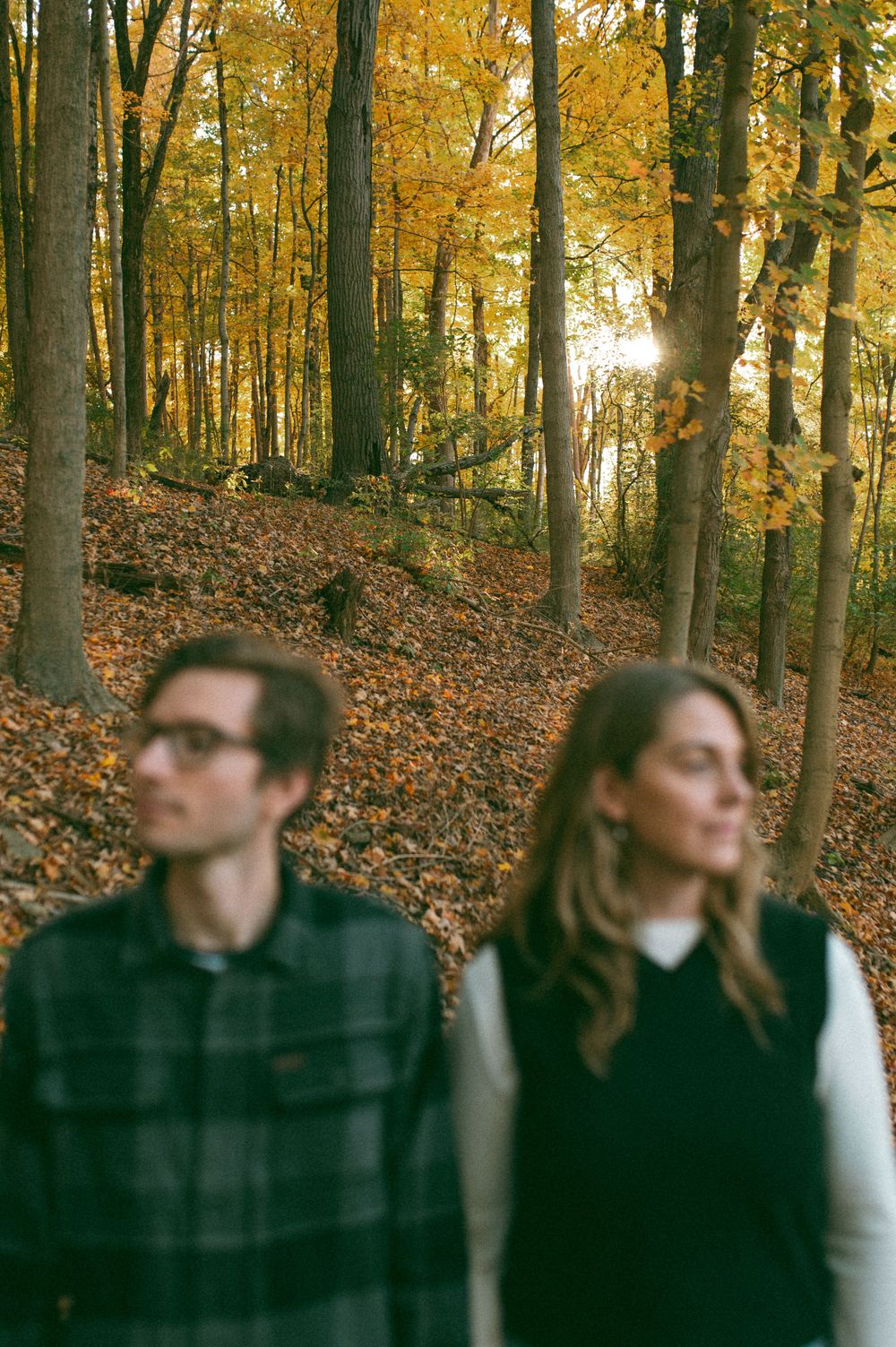 Stylish couple posing separately in a fall forest with warm sunlight filtering through trees.