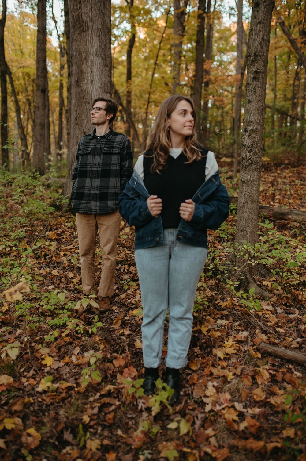 Two people looking off in different directions while standing among fall foliage in a forest.