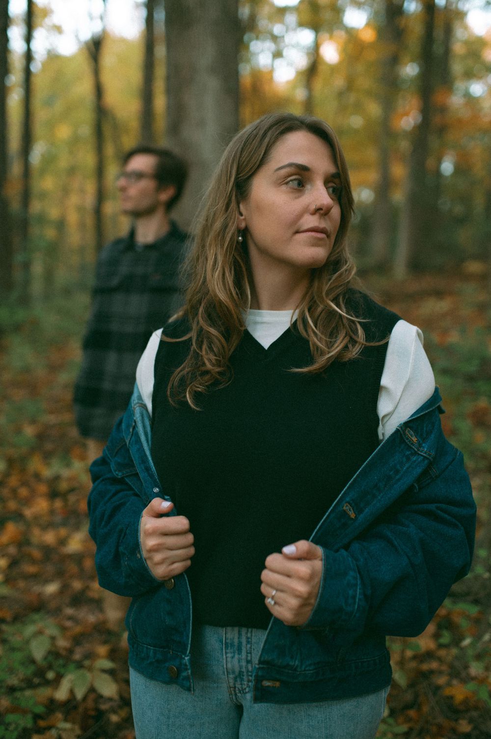 Person in blue denim jacket and black vest stands in foreground of autumn forest scene.