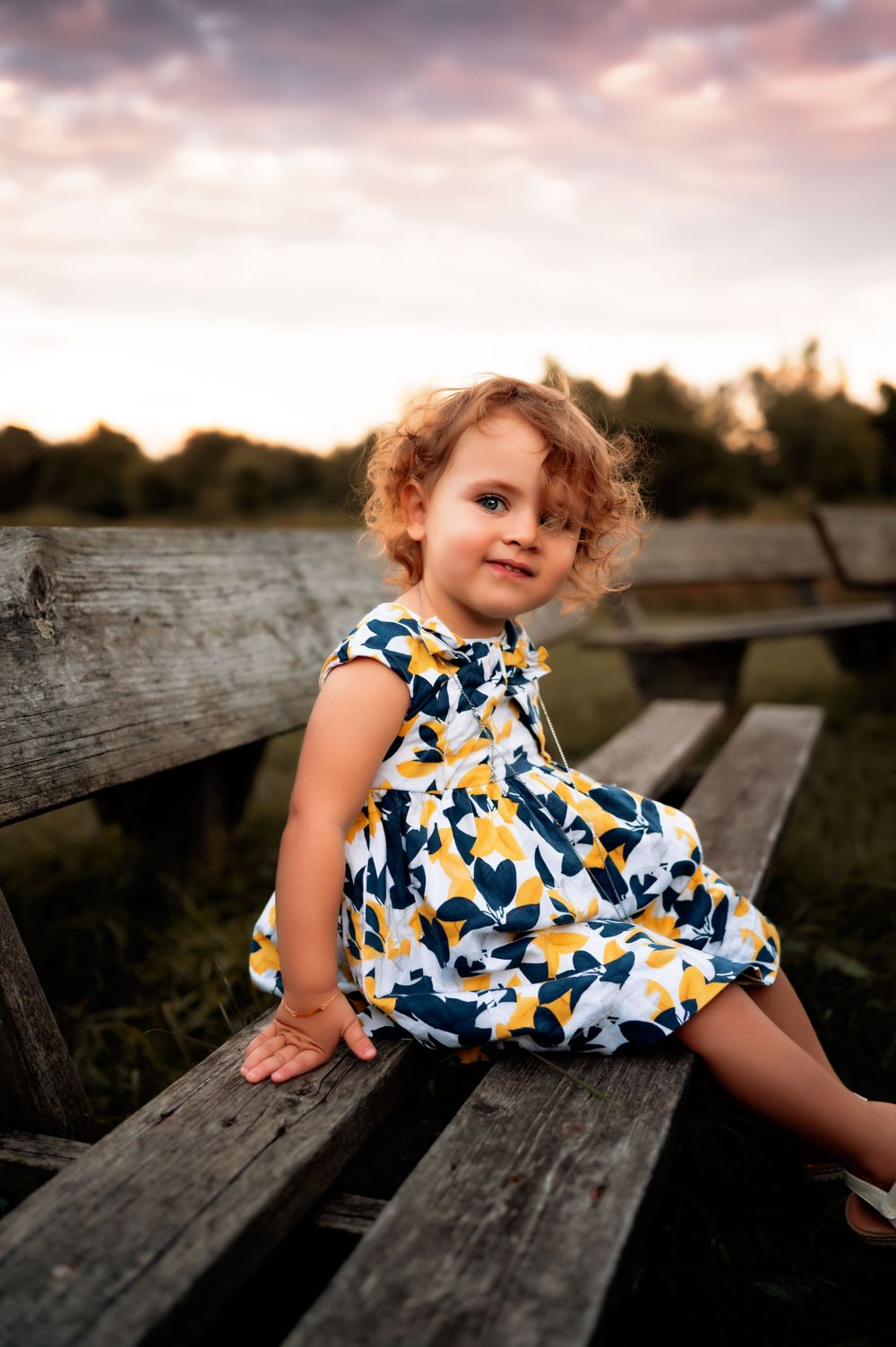 Young child in a blue and yellow floral dress sits on wooden bleachers at sunset.