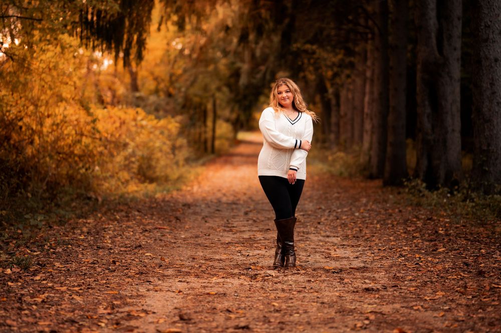 Person wearing white sweater walks along a tree-lined autumn path during golden hour.