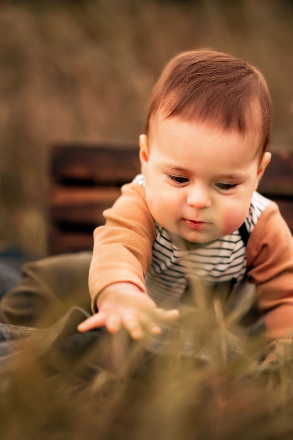 Baby in striped outfit reaching toward grass while exploring outdoors.