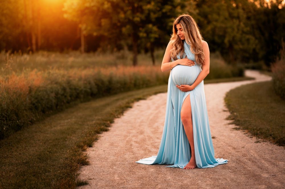 Expectant mother in flowing blue dress poses on dirt path during golden sunset maternity photoshoot.