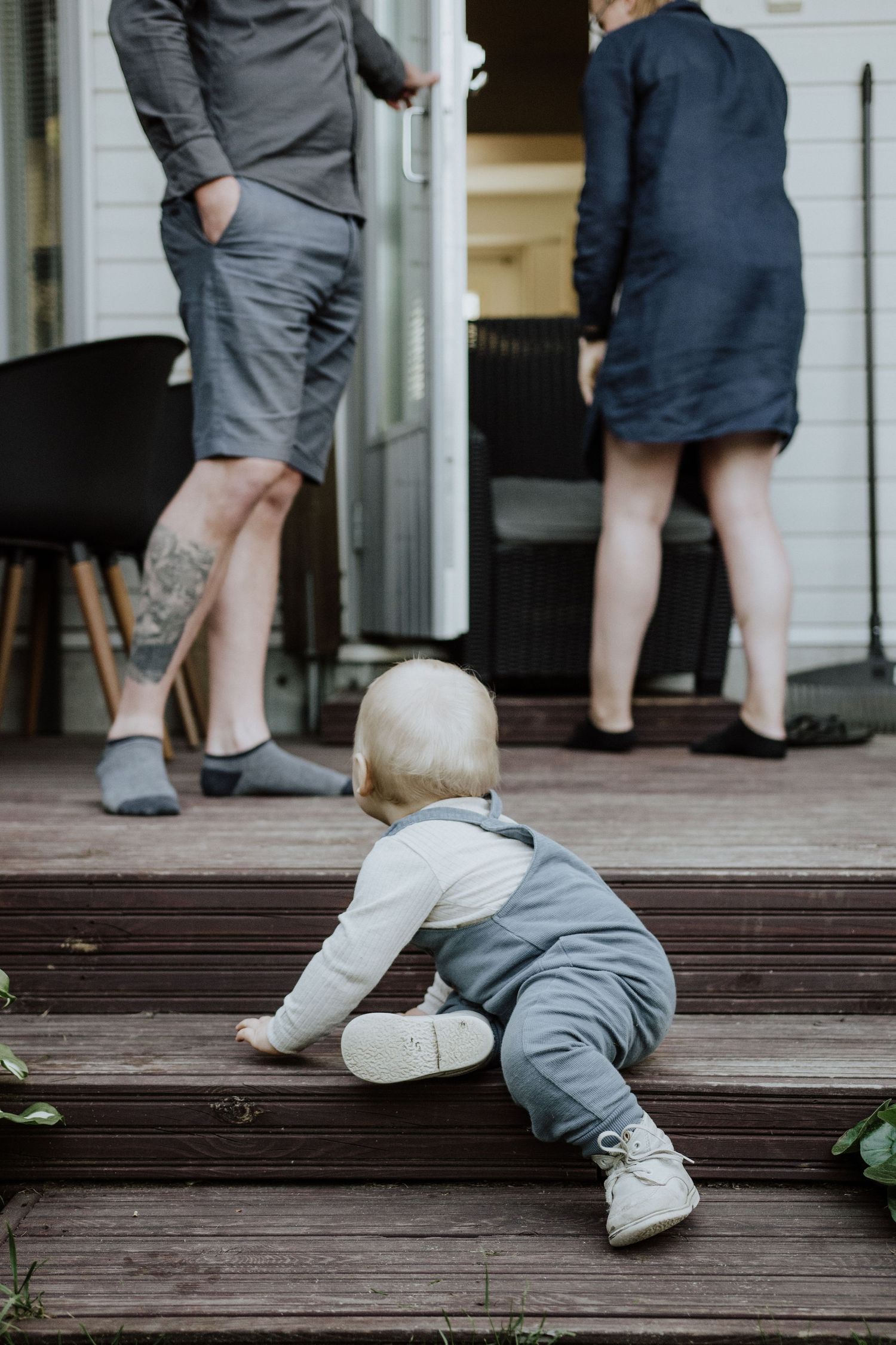 A baby crawls on wooden deck steps while adults stand nearby.