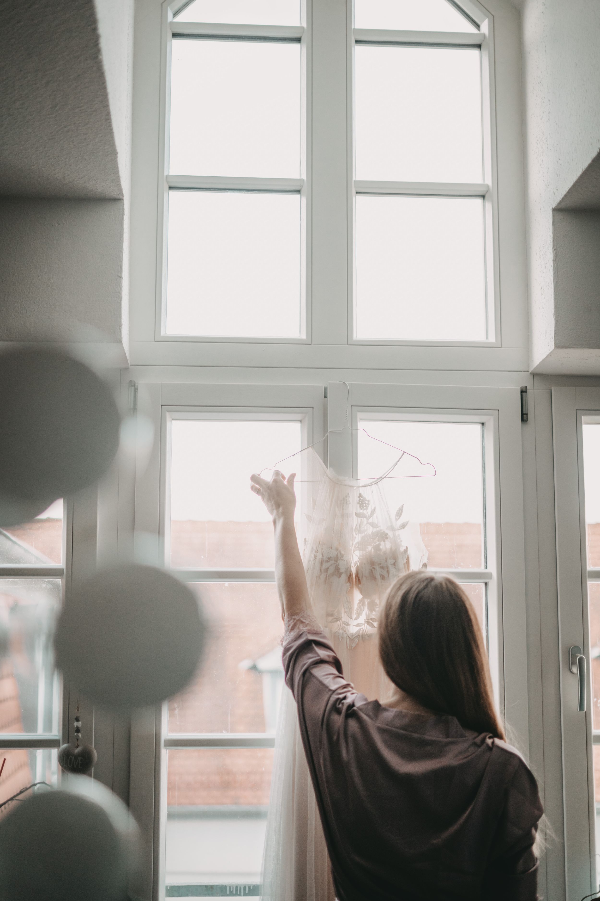 Person hanging wedding dress near tall arched window with soft natural light.